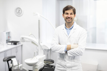 Naklejka na ściany i meble Dental Center. Portrait of male dentist doctor posing at workplace, stomatologist standing with folded arms in modern clinic interior