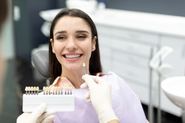 Cheerful young female patient and professional dentist choosing filling shade, using tooth scale...