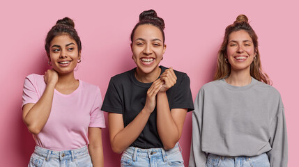 Photo of three diverse young women express positive emotions smile broadly hear pleasant news dressed in casual clothes and jeans isolated over pink background. People and positiveness concept - obrazy, fototapety, plakaty