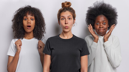 Photo of shocked European woman with hair bun wears casual black t shirt poses between two African women with curly hair feel impressed isolated over white background. Collage shot. Human reactions
