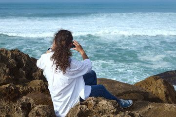 Fototapeta na wymiar Curly haired multi ethnic pretty woman taking photo on her smart mobile phone, sitting on the rock on the Atlantic ocean beach