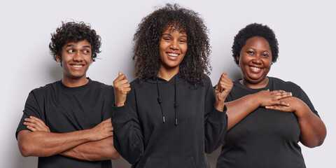 Photo of cheerful curly haired young female model clenches fists celebrates success smiling Hindu man keeps arms crossed and focused aside thankful African American woman makes gratitude gesture