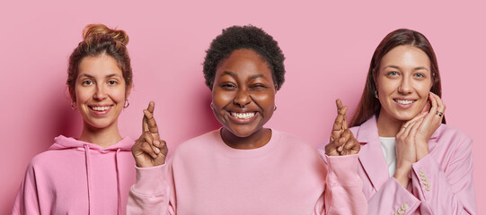 Studio shot of three women stand next to each other smile happily keep fingers crossed believe in good luck express positive emotions isolated over pink background. People and body language concept - obrazy, fototapety, plakaty