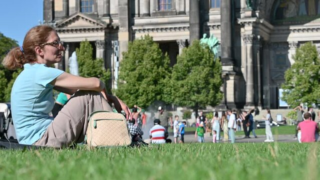 Berlin, Germany, August 10, 2023. A middle-aged woman enjoys some relaxation on the lawn in front of the cathedral, in the blurred background other people and the facade. Beautiful summer day.