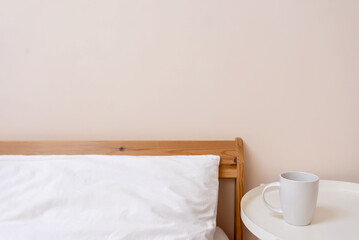 Fototapeta na wymiar Lifestyle minimal bedroom interior detail, bed with white pillow. coffee cup on bedside table, empty peachy neutral wall background