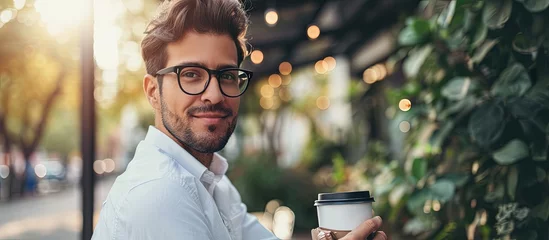 Fototapeten Professional modern and confident young businessman with glasses and white shirt holding a eco friendly paper cup of coffee in urban park of business district. Creative Banner. Copyspace image © HN Works