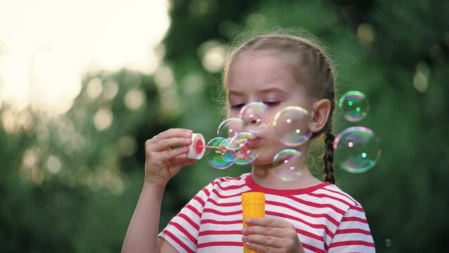 Joyful little girl with thin braids blows soap bubbles in surrounding of blurry garden. Colorful soap bubbles flying around carelessly under gaze of little girl. Girl spends free time blowing bubbles