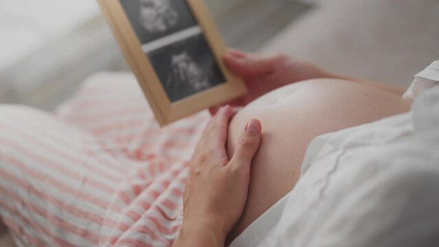 A pregnant girl in loungewear looks at a photo of her unborn child's bridles. White woman holds in her hands a frame with a photo of the reins caring for the future of mother and baby, child