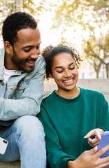 Vertical shot of two young latin american people using smart phone together sitting outdoors. Cheerful african american friends laughing while looking at cellphone screen. Social media concept.