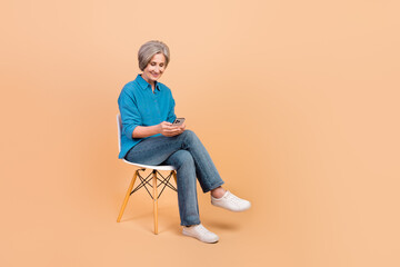 Full size photo of good mood smart woman dressed stylish shirt sit on chair look at smartphone isolated on pastel color background