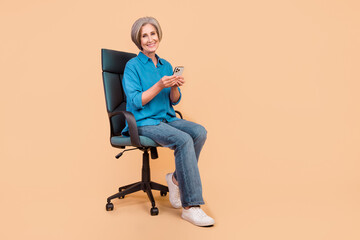 Full size photo of smart pleasant woman dressed stylish shirt sit on armchair with smartphone in hands isolated on pastel color background