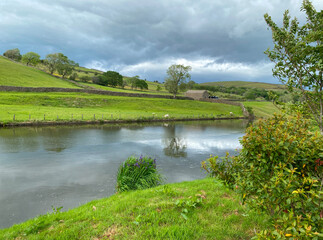 View over the village dam, that supplied water to the mills, with grass, wild plants, and distant hills in, Lothersdale, UK