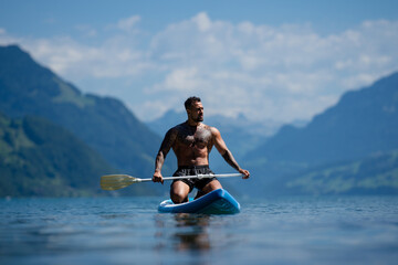 Man on paddle board in Alps lake mountains. Leisure activities with paddle on Lake in Switzerland. Man paddle surfing board on Geneva Lake. Muscular sexy strong Hispanic man paddle board surfers.