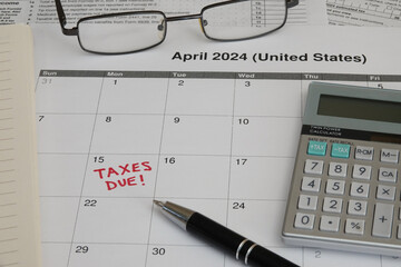 A 2024 calendar noting the April 15 USA Internal Revenue Service IRS income filing deadline for year 2023 taxes is shown up close, with a calculator, ink pen, notebook, and glasses in the frame.