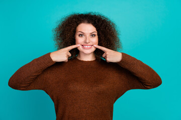 Photo of young lady in brown jumper directing fingers to white toothy smile after stomatology...