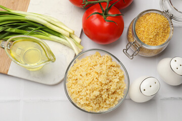 Delicious bulgur in bowl, vegetables, oil and spices on white tiled table, flat lay