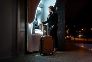 Attractive business woman at ATM with luggage at night , withdrawing funds 