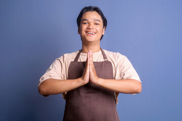 Young male barista in brown apron showing greeting and welcoming gesture with happy expression