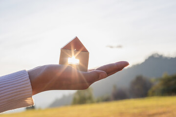 Woman hand holds wooden house against the sun. - obrazy, fototapety, plakaty