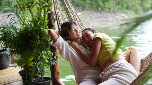 4K Happy Asian LGBTQ couple relaxing on hammock together at lake house balcony. People enjoy and fun outdoor lifestyle travel nature on summer holiday vacation. LGBT person and pride day concept.
