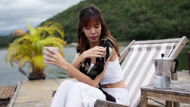 Young Asian woman relax and enjoy outdoor lifestyle travel nature forest mountain on summer holiday vacation. Generation z girl resting on lake house balcony and drinking coffee and reading a book.