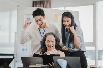 Asian office worker discussing with younger colleagues over laptops in a meeting room, possibly reviewing monthly reports or sharing opinions.