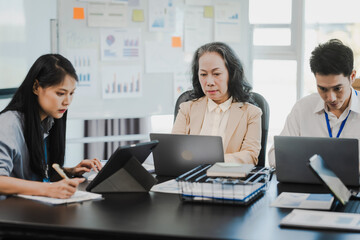 Asian office worker discussing with younger colleagues over laptops in a meeting room, possibly reviewing monthly reports or sharing opinions.