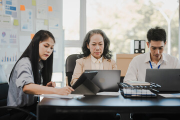 Asian office worker discussing with younger colleagues over laptops in a meeting room, possibly reviewing monthly reports or sharing opinions.