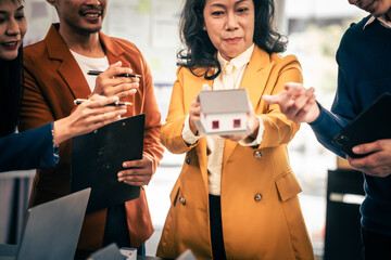 Asian real estate team engaged in a discussion, with two men and a woman focusing on a house model on a table, suggesting a planning or sales meeting.