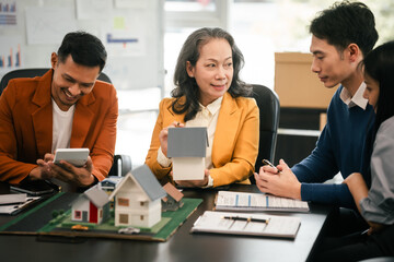 Asian real estate team engaged in a discussion, with two men and a woman focusing on a house model on a table, suggesting a planning or sales meeting.