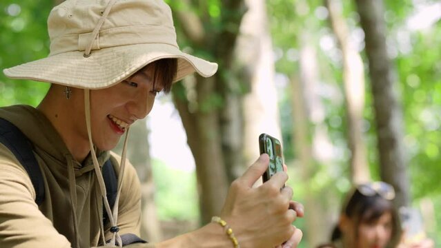 Young Asian man and woman friends travel nature on summer holiday vacation. Generation z people enjoy and fun outdoor lifestyle using smartphone taking picture of beautiful nature at forest mountain.