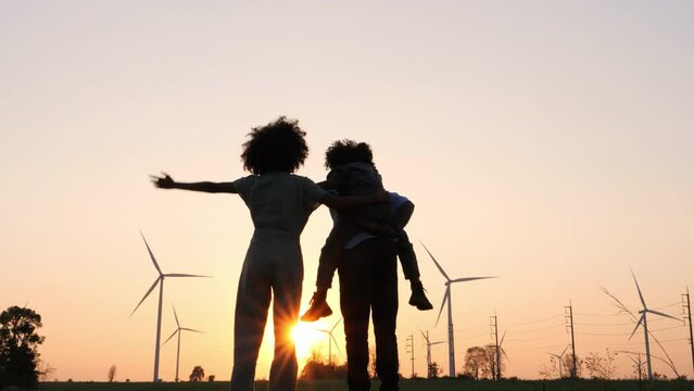 Silhouettes Of Happy Family Father, Mother And Child Daughter Sits On The Shoulders Of His Father With Windmills For Electricity Generation At Sunrise.	