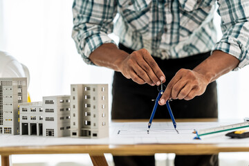 Close up Asian individual architect or engineer holding building scale model of multi-story building. design or review of architectural plans laid out on work surface in front of them.