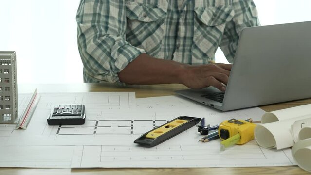 Close up Asian individual architect or engineer holding building scale model of multi-story building. design or review of architectural plans laid out on work surface in front of them.