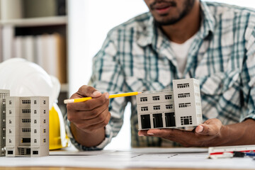 Close up Asian individual architect or engineer holding building scale model of multi-story building. design or review of architectural plans laid out on work surface in front of them.