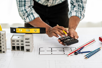 Close up Asian individual architect or engineer holding building scale model of multi-story building. design or review of architectural plans laid out on work surface in front of them.
