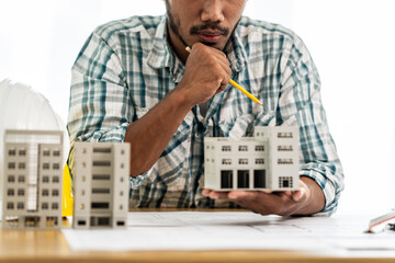 Close up Asian individual architect or engineer holding building scale model of multi-story building. design or review of architectural plans laid out on work surface in front of them.