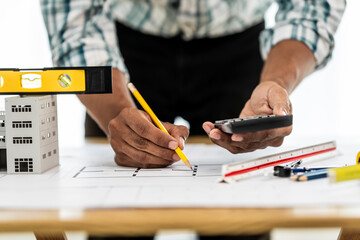 Close up Asian individual architect or engineer holding building scale model of multi-story building. design or review of architectural plans laid out on work surface in front of them.