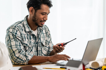 Asian person hands working on architectural blueprints with a pencil, alongside scale models of houses, a laptop, and construction tools on the desk.