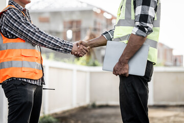 two construction workers, one Asian, shaking hands on a building site, possibly indicating...