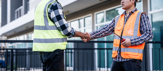 two construction workers, one Asian, shaking hands on a building site, possibly indicating agreement or completion of a task, with a modern building in the background. - obrazy, fototapety, plakaty