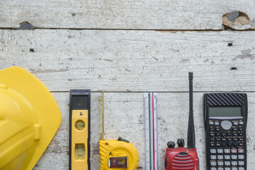 Top view construction tools such as a yellow hard hat, spirit level, measuring tape, folding ruler arrayed against a wooden plank background.