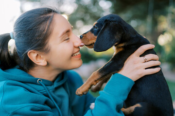 Black puppy licks the nose of a smiling girl holding him in her arms - obrazy, fototapety, plakaty