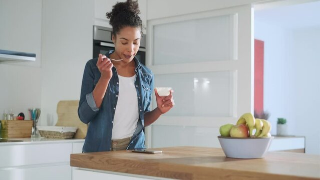 Smiling woman eating yoghurt and checking text messages in kitchen