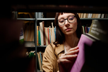 Young student looking for books on shelf in library