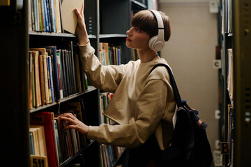 Student wearing wireless headphones and taking book from shelf in library