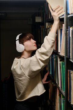 Young student wearing wireless headphones and taking book from top shelf in library