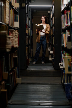 Young student reading book amidst shelves in library