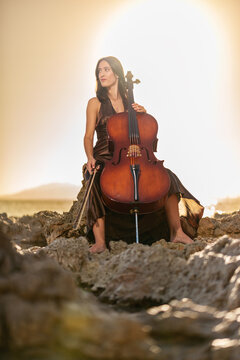 Woman sitting on the rocks on the shore with a lost look holding the chello at sunset