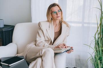 Professional woman in a beige suit thoughtfully reading a book in a well-lit room, with sheer...
